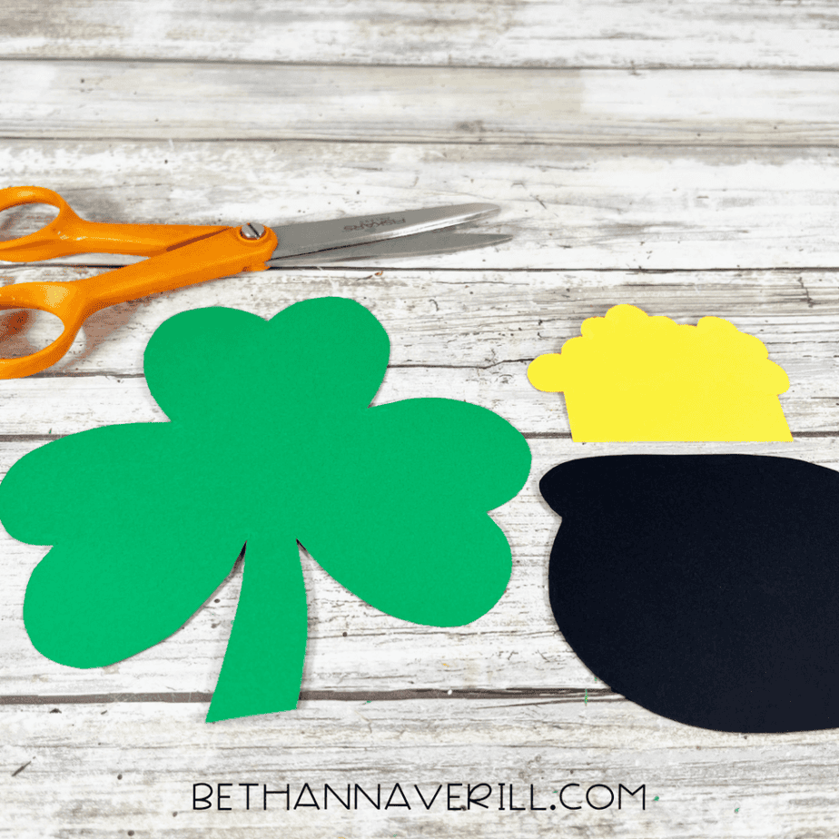 Cut out cardstock shapes including a green shamrock, a black pot, and a yellow cloud shape placed on a wooden surface with scissors nearby.