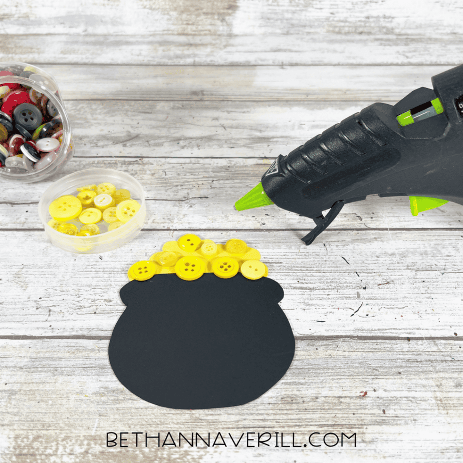A black cardstock pot shape decorated with yellow buttons along the top edge. A hot glue gun and containers of buttons are visible on the wooden surface.