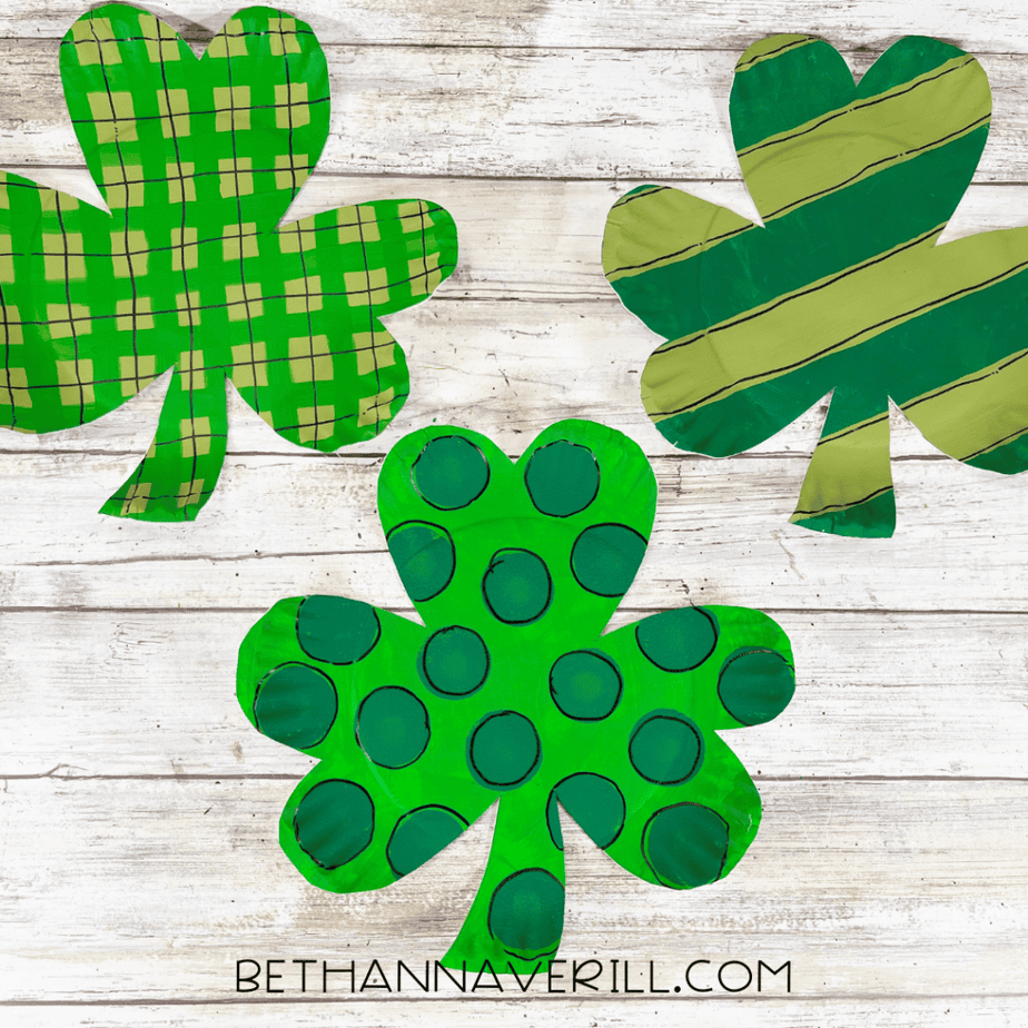 Three completed shamrock paper plate crafts displayed on a wood background.
