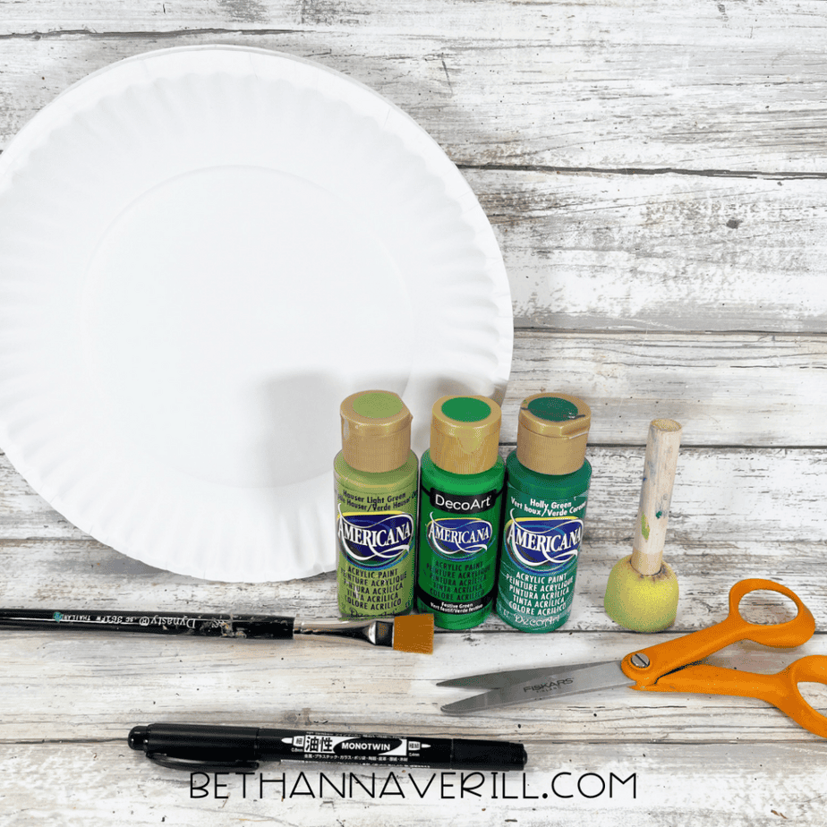 White paper plate with three bottles of green acrylic paint, a paintbrush, foam dauber, scissors, and a black marker on a wood background.