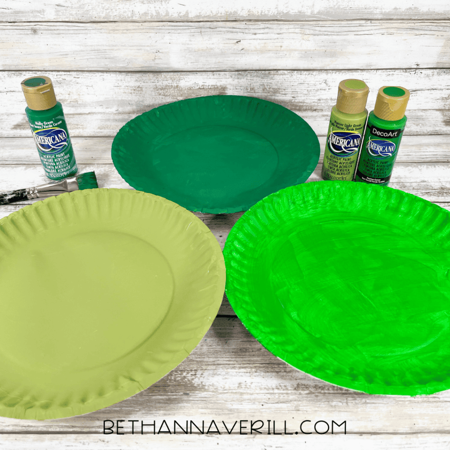Three paper plates painted in different shades of green with paint bottles and a paintbrush displayed beside them.