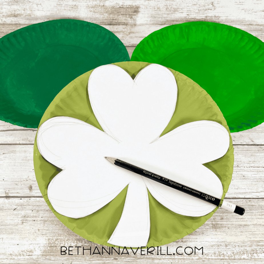 Paper shamrock template placed on a light green painted paper plate with a pencil resting on top.