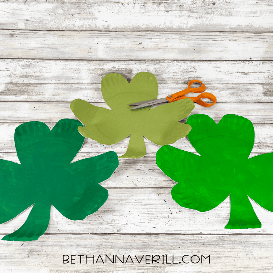 Three shamrock shapes cut from painted paper plates with orange scissors on a wood surface.