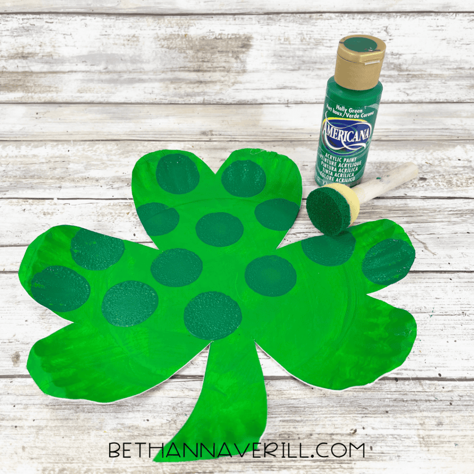 Green shamrock cutout with dark green painted circles and a foam dauber beside a bottle of acrylic paint.