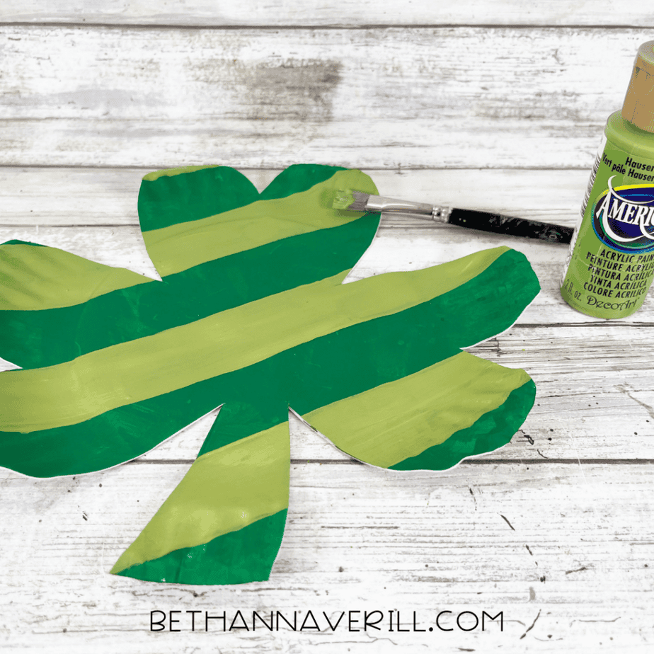 Shamrock cutout painted with alternating light and dark green stripes and a paintbrush next to a bottle of paint.