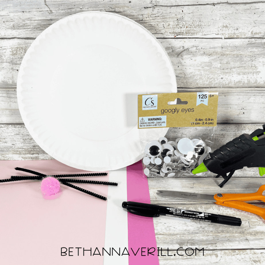 Craft supplies for a bunny paper plate craft including a white paper plate, googly eyes, pipe cleaners, pink and white paper, scissors, a glue gun, and a black marker on a wood-patterned surface.