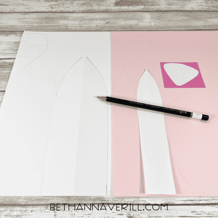 White and pink paper with traced bunny ear shapes, a pencil, and cut-out paper pieces on a wood-patterned surface.
