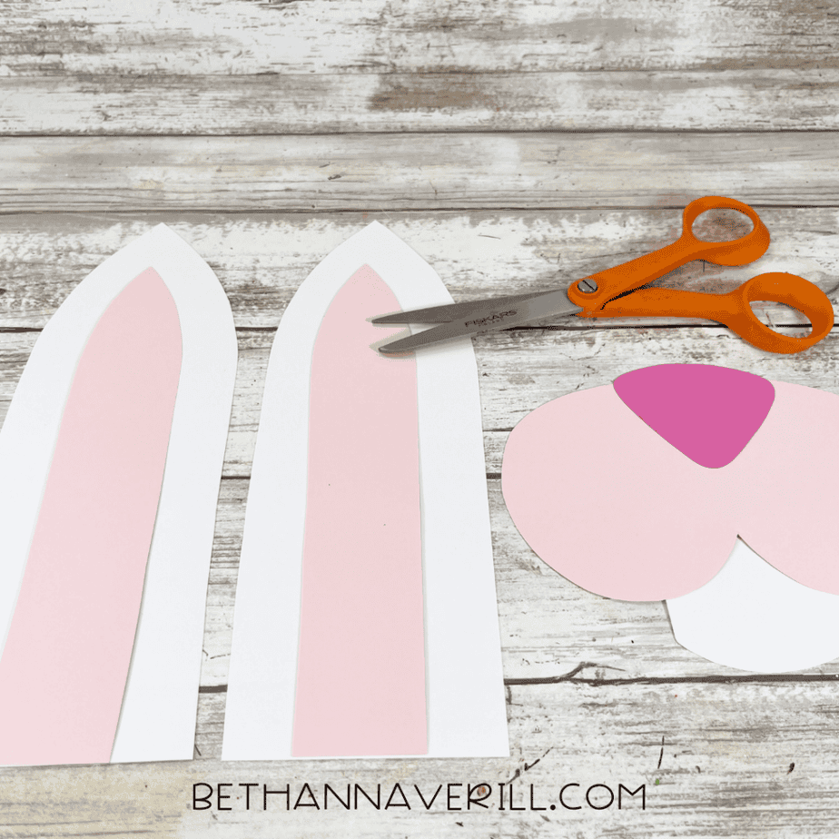 Two paper bunny ears, orange-handled scissors, and cut-out paper pieces for a bunny face on a wood-patterned surface.