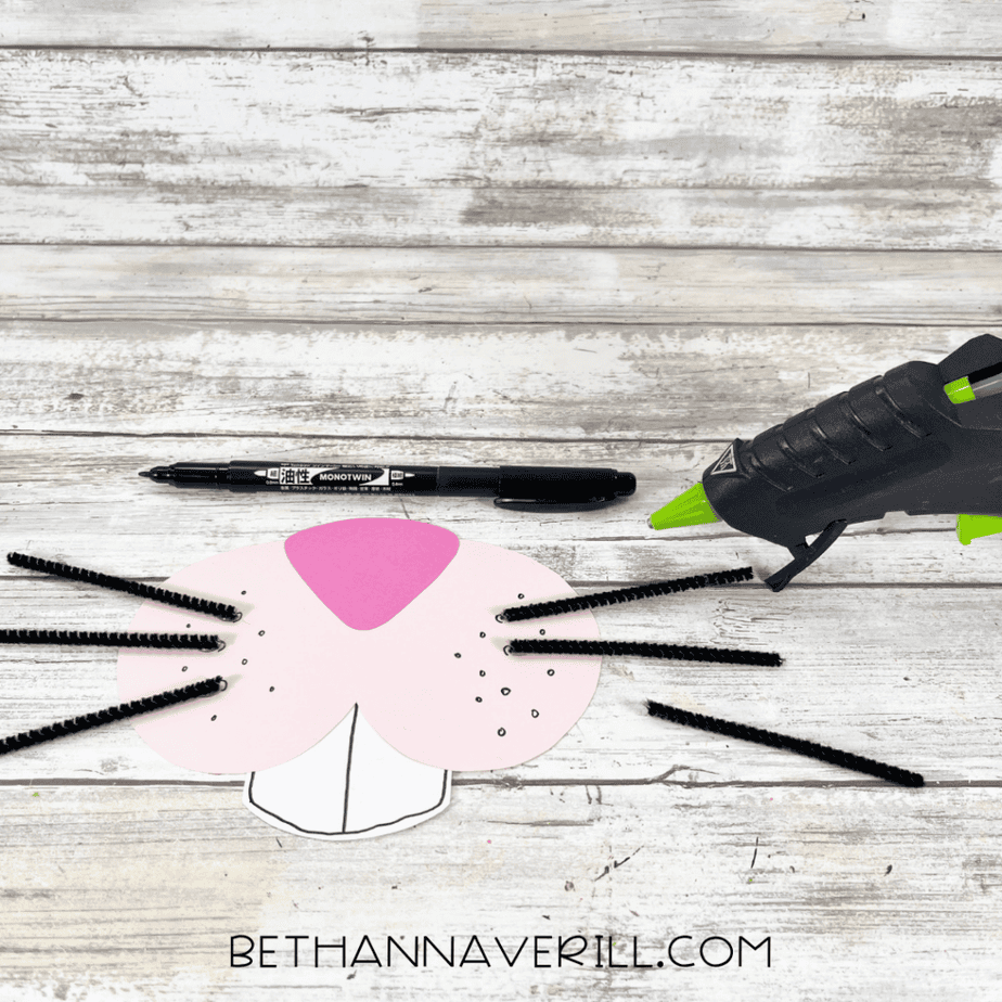 Paper bunny face pieces with black pipe cleaner whiskers attached, displayed beside a marker and hot glue gun.