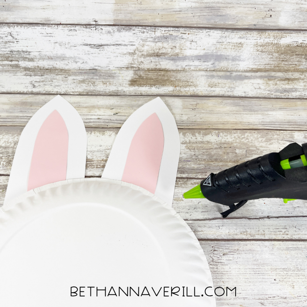 A white paper plate with attached paper bunny ears beside a hot glue gun on a wood-patterned surface.