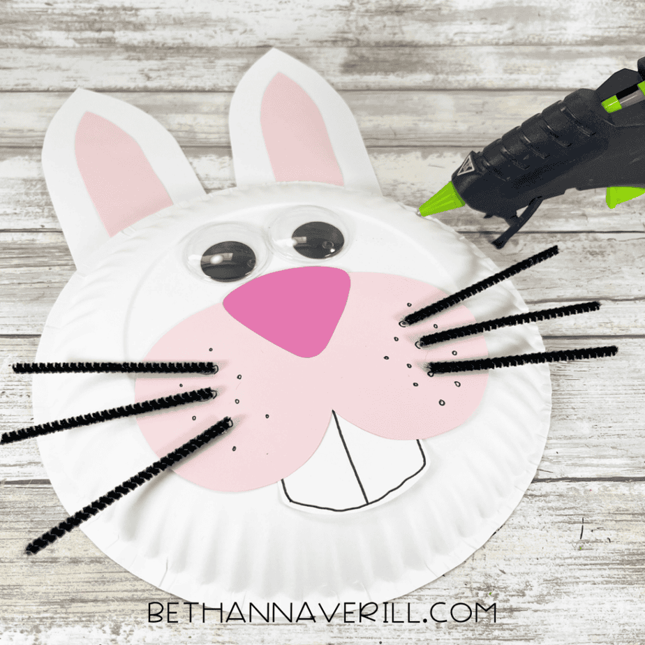 A partially assembled bunny paper plate craft with googly eyes, whiskers, pink cheeks, and buck teeth attached to the plate.