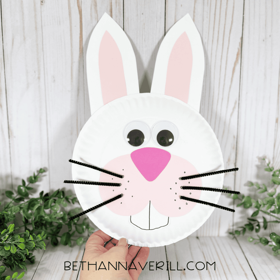 A hand holding a completed bunny paper plate craft with large ears, googly eyes, whiskers, pink cheeks, and buck teeth in front of a wood-patterned background with greenery.