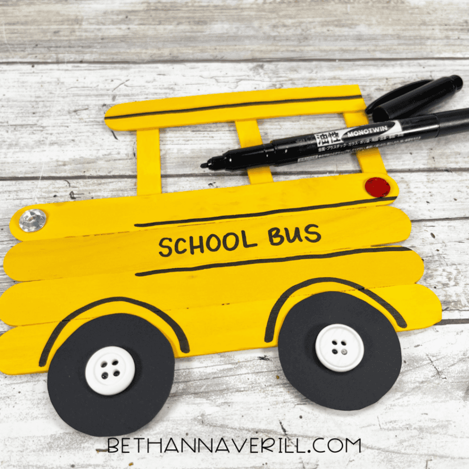 Marker drawing details on a yellow popsicle stick school bus craft, including lines and the words “school bus.”