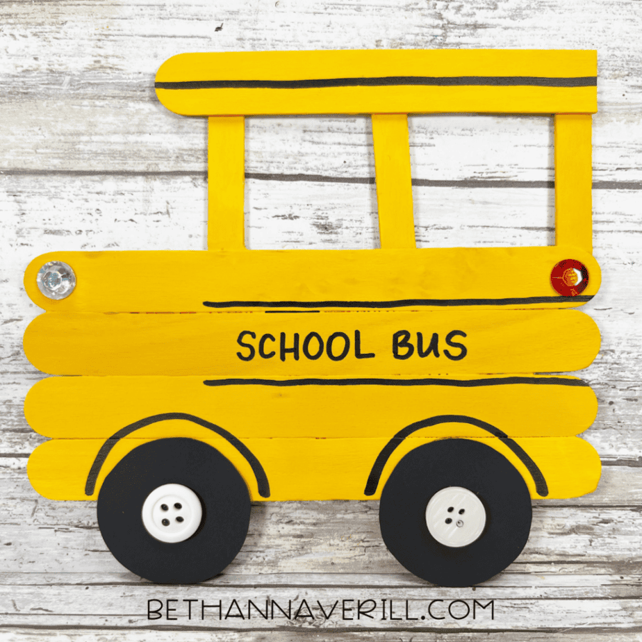 Finished popsicle stick school bus craft painted yellow with black wheels and button centers on a wooden background.