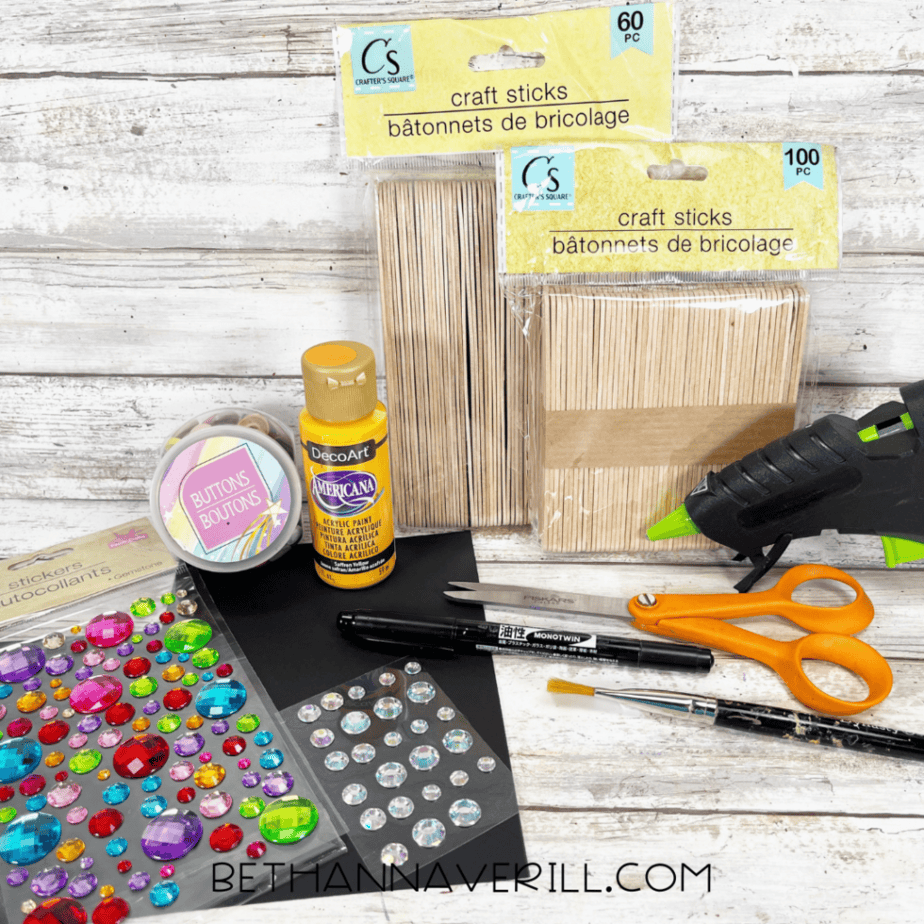 Craft supplies for a popsicle stick craft including wooden craft sticks, acrylic paint, glue gun, scissors, buttons, rhinestone stickers, paintbrush, and black marker on a wooden background.