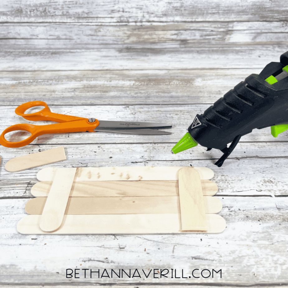Wooden craft sticks glued together in a rectangle shape while making a popsicle stick craft, with scissors and a hot glue gun on a wooden table.