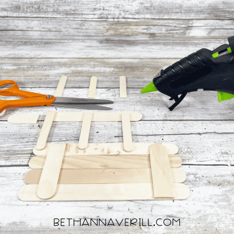 Additional craft sticks being glued onto a popsicle stick base to create a bus-shaped frame, with scissors and a hot glue gun nearby.