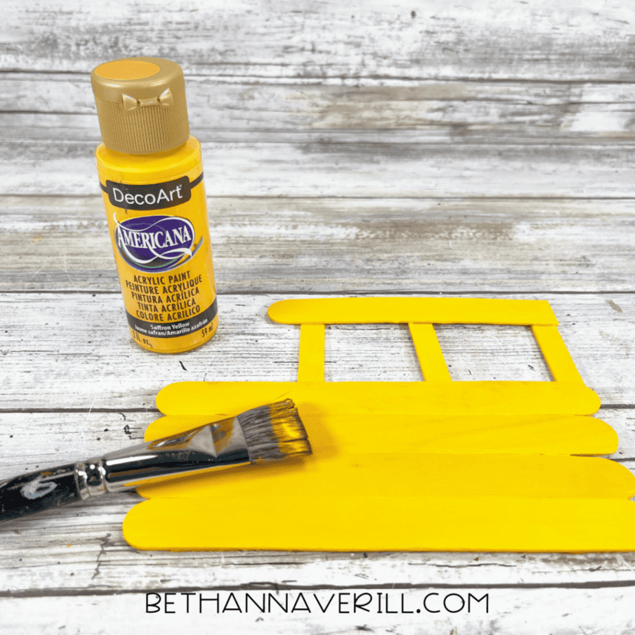 Yellow acrylic paint being brushed onto a popsicle stick craft base, covering the wooden sticks completely.