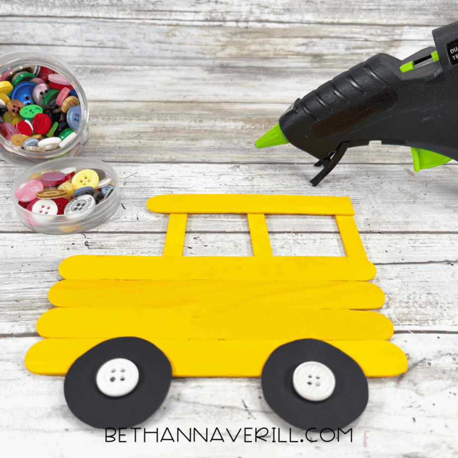 Painted yellow popsicle stick bus craft with black cardstock wheels and white buttons attached in the center as wheel hubs.