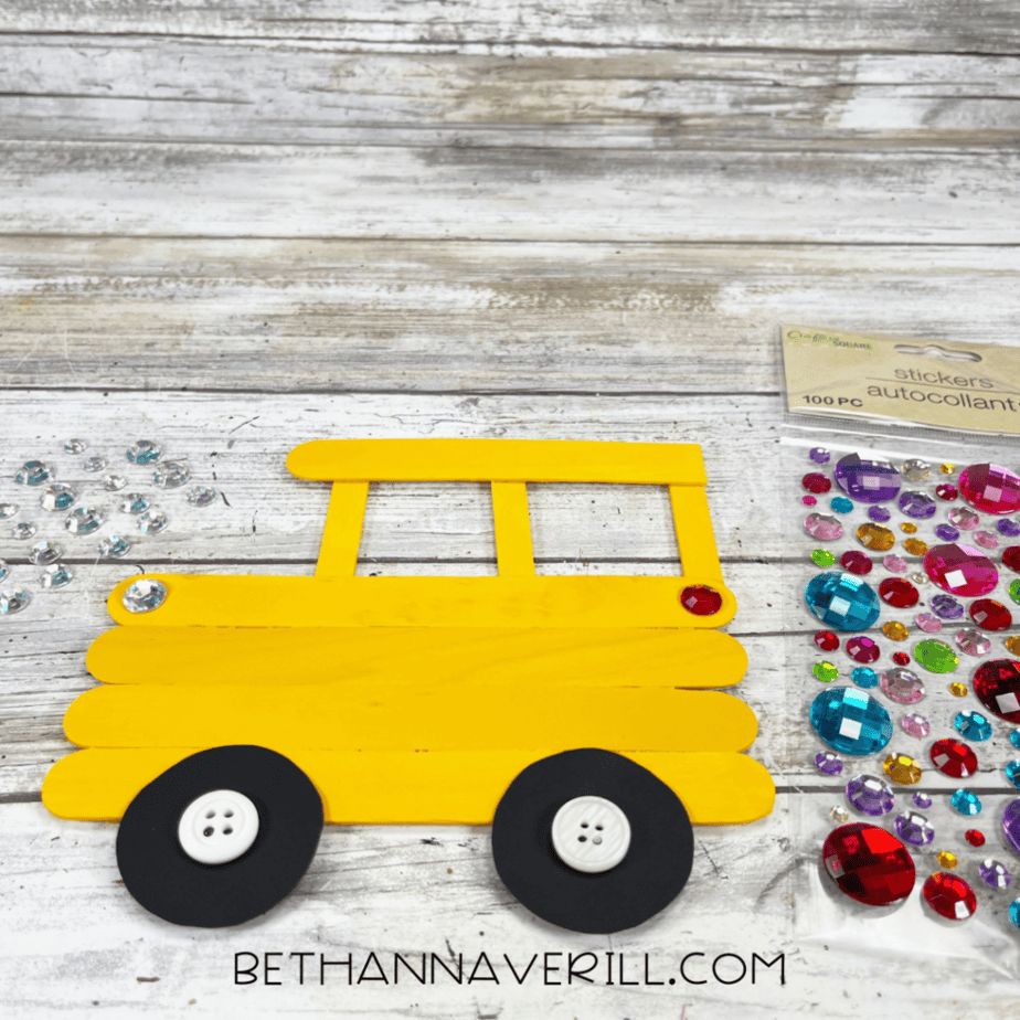 Yellow popsicle stick bus craft decorated with rhinestone stickers and black cardstock wheels on a wooden background.