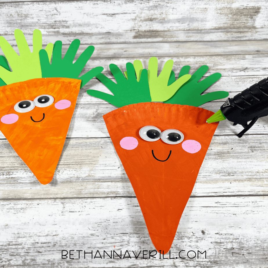 Green and light green hand-shaped paper pieces being attached to the top of an orange triangular paper plate piece using a hot glue gun.