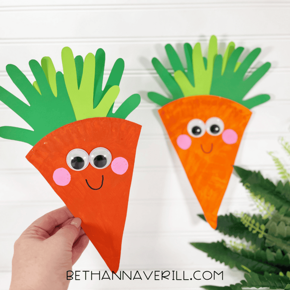 A hand holding a finished carrot craft made from an orange triangular paper plate piece with googly eyes, pink cheeks, a drawn smile, and layered green hand-shaped paper pieces at the top, with another finished carrot craft in the background.