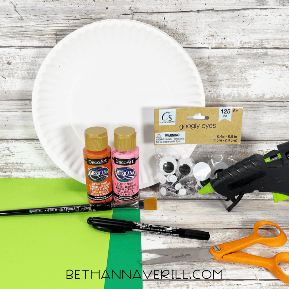 Craft supplies arranged on a table including a white paper plate, two bottles of acrylic paint, a paintbrush, a black marker, googly eyes in packaging, a hot glue gun, scissors, and green paper.