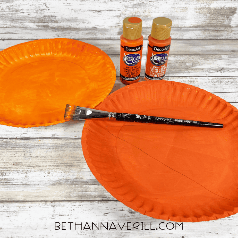Two paper plates painted orange with a paintbrush resting across one plate and two bottles of orange acrylic paint nearby.