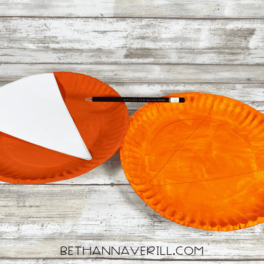 Two orange painted paper plates with pencil lines drawn on them and a white triangular template placed on one plate.