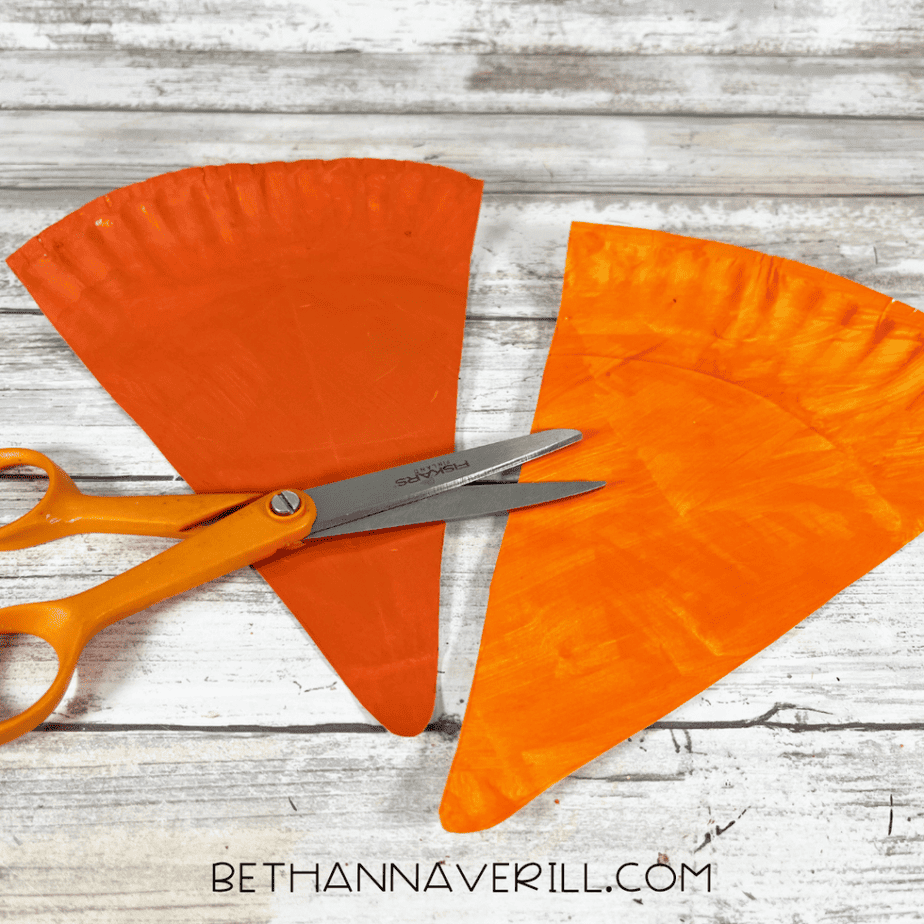 Scissors cutting two orange triangular pieces from painted paper plates.