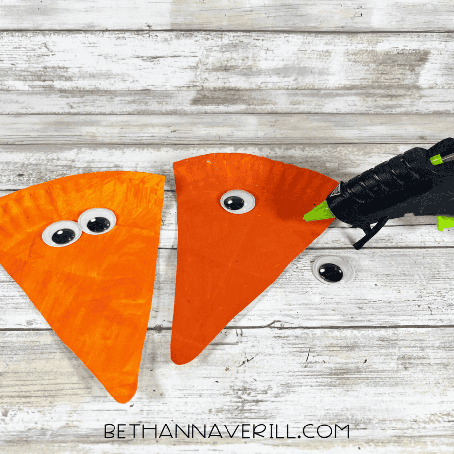 Two orange triangular paper plate pieces with googly eyes being attached using a hot glue gun.