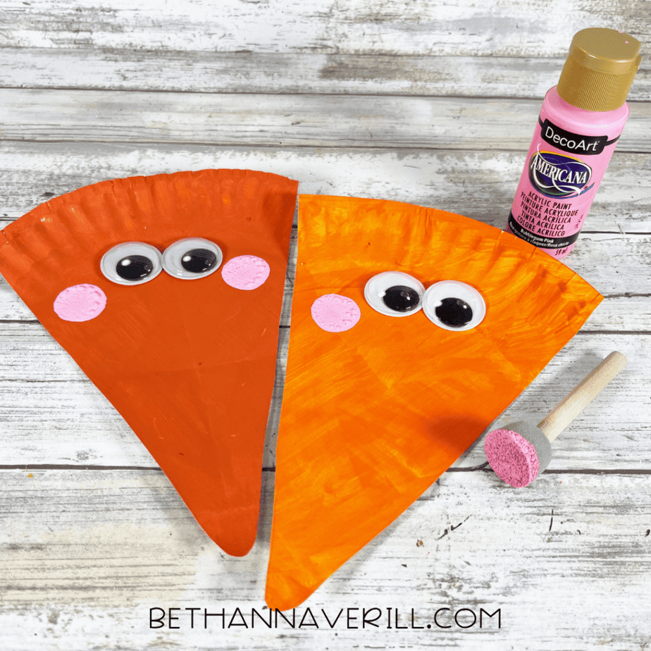 Two orange triangular paper plate pieces with googly eyes and pink painted circles on each side of the eyes, with a pink paint bottle and a foam dauber nearby.