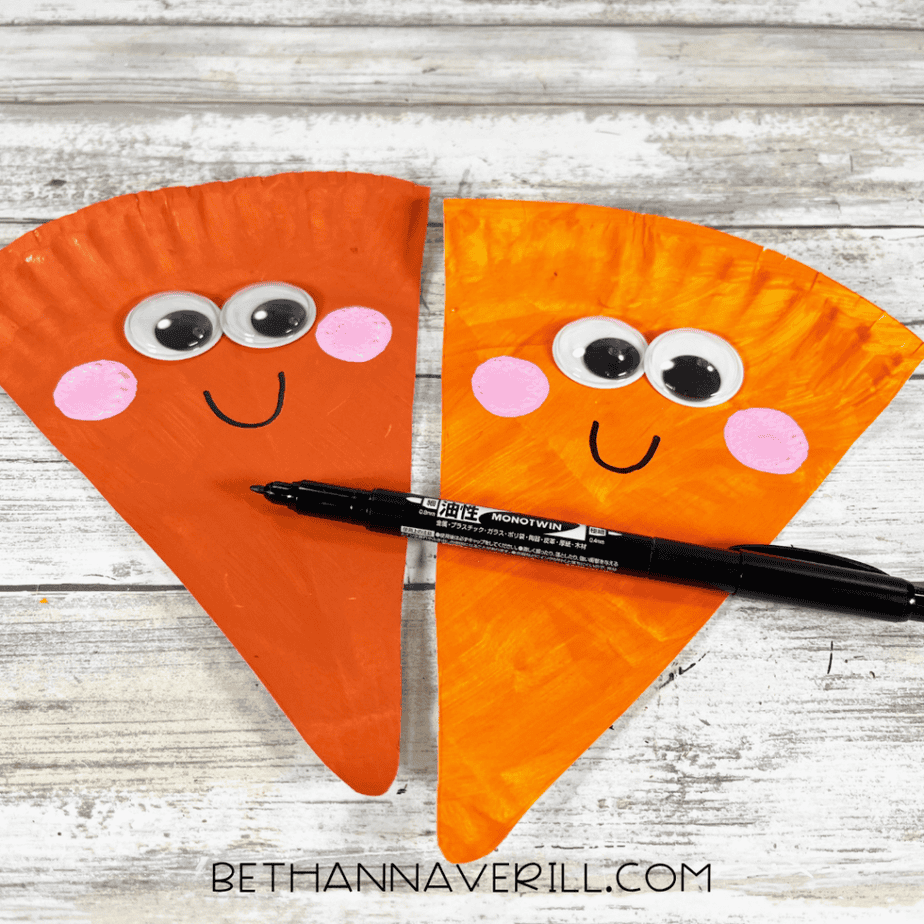 Two orange triangular paper plate pieces with googly eyes and pink cheeks while a black marker draws a curved smile.