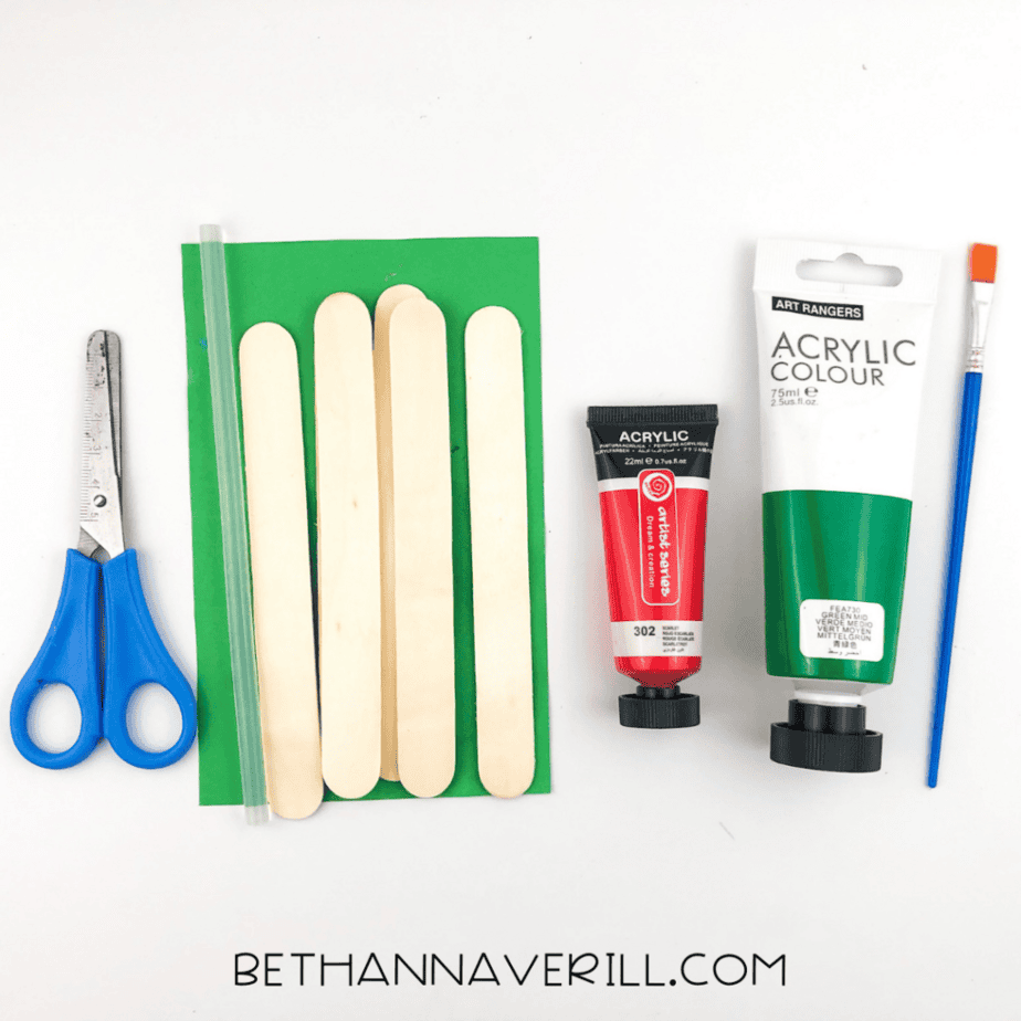 Craft supplies on a white background including scissors, a glue stick, wooden craft sticks, green paper, red and green acrylic paint tubes, and a paintbrush.