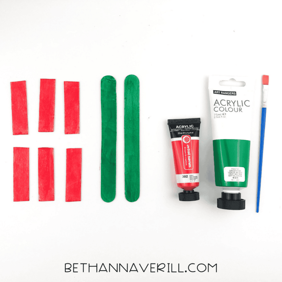 Six wooden craft sticks painted red and two wooden craft sticks painted green, with paint tubes and a paintbrush beside them.