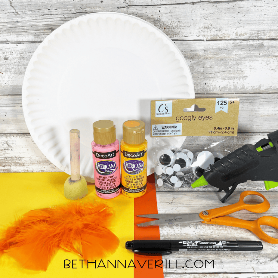 Craft supplies arranged on a table including a white paper plate, yellow and pink acrylic paint bottles, a foam dauber, googly eyes, a hot glue gun, scissors, orange feathers, colored paper, and a black marker.