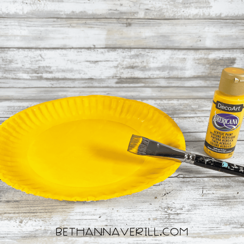 A paper plate being painted yellow with a paintbrush next to a bottle of yellow acrylic paint.