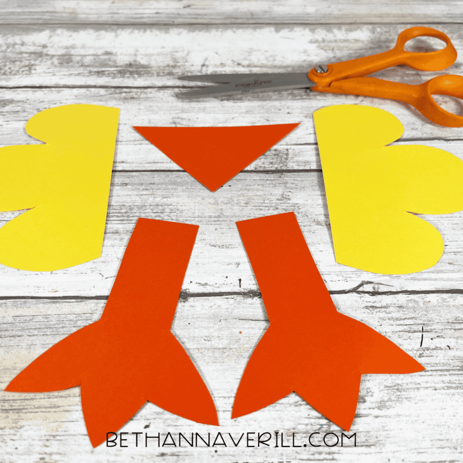 Cut paper pieces arranged on a table including two orange feet shapes, a triangle beak, and two yellow wing shapes, with scissors nearby.