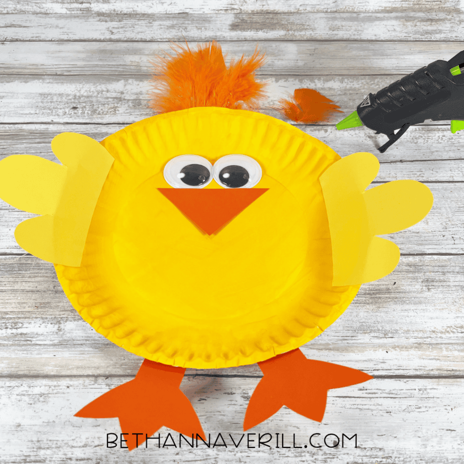 Orange feathers being attached to the top of a yellow paper plate chick craft that has eyes, a beak, wings, and feet.