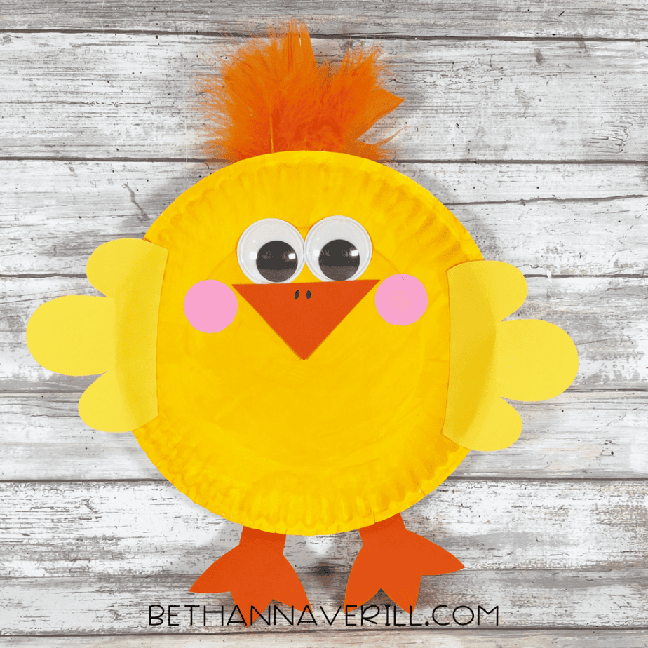 The completed chick craft made from a yellow paper plate with googly eyes, a triangular beak, pink cheeks, yellow wings, orange feet, and orange feathers on top.