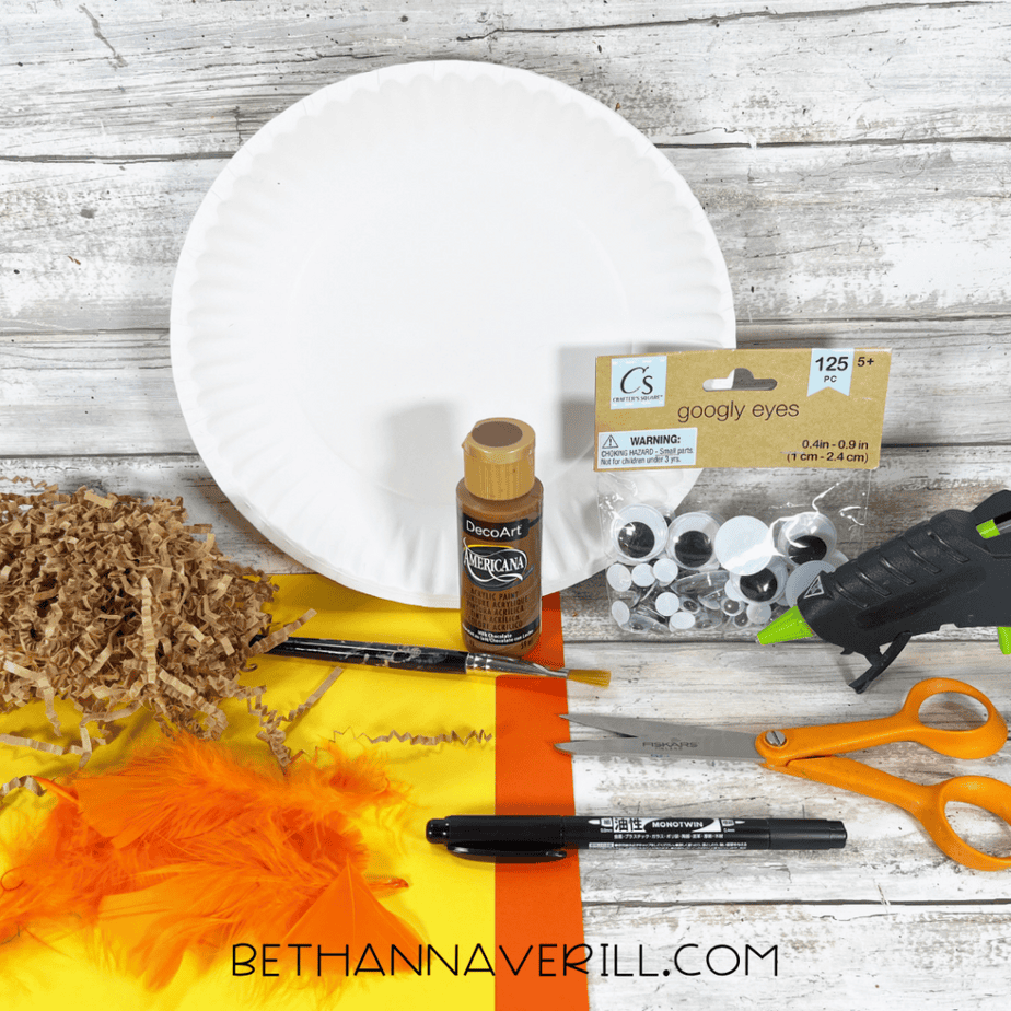 A white paper plate sits on a wooden surface. Around it are craft materials: a small bottle of brown paint, a paintbrush, a bag of googly eyes, a hot glue gun, scissors, a black marker, sheets of yellow and orange paper, shredded brown paper filler, and orange feathers.