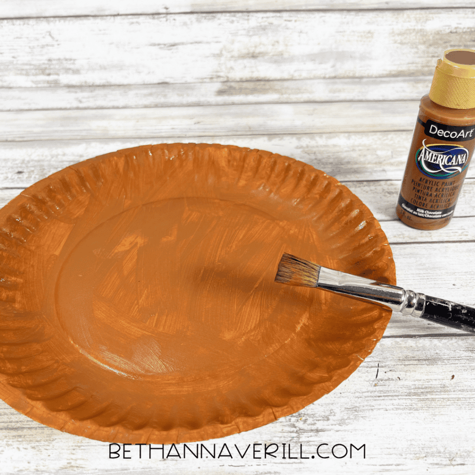 The paper plate is fully painted brown. A paintbrush rests on the plate, and the paint bottle is nearby.
