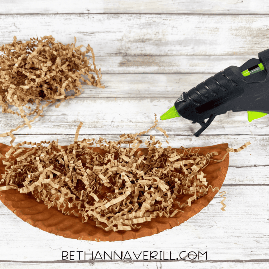 One half of the plate has shredded brown paper glued onto it. A hot glue gun is positioned nearby, and extra shredded paper sits off to the side.