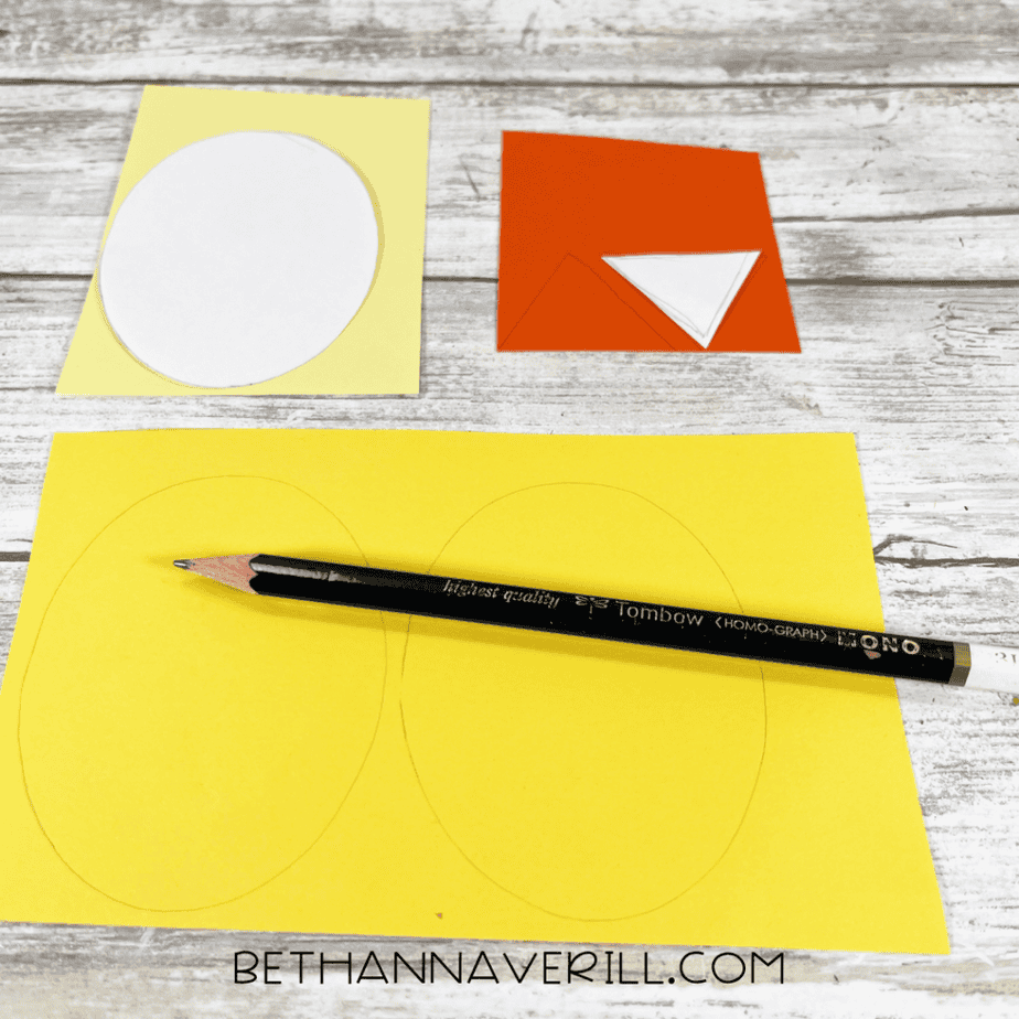 Paper shapes are prepared: yellow oval shapes and orange triangle shapes are drawn or cut from colored paper. A pencil and scissors are visible.