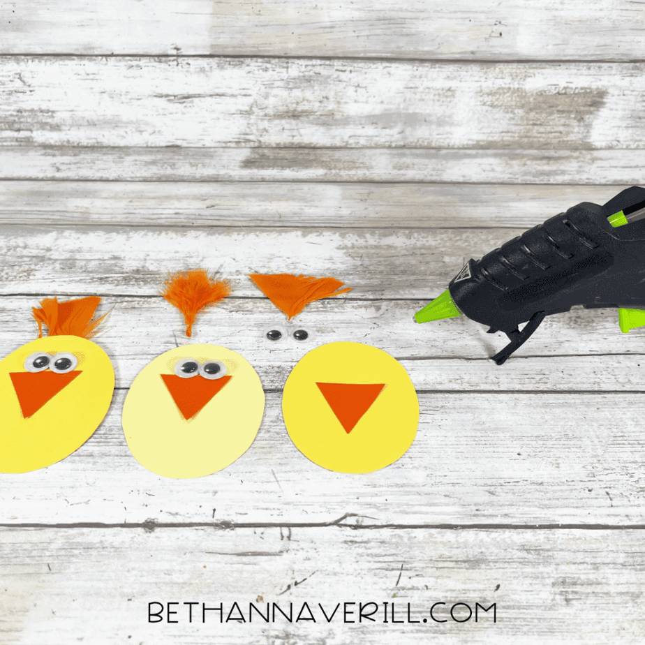 The yellow ovals now have added elements: googly eyes, orange triangle shapes attached, and small orange feather pieces placed on top. A hot glue gun is visible to the right.
