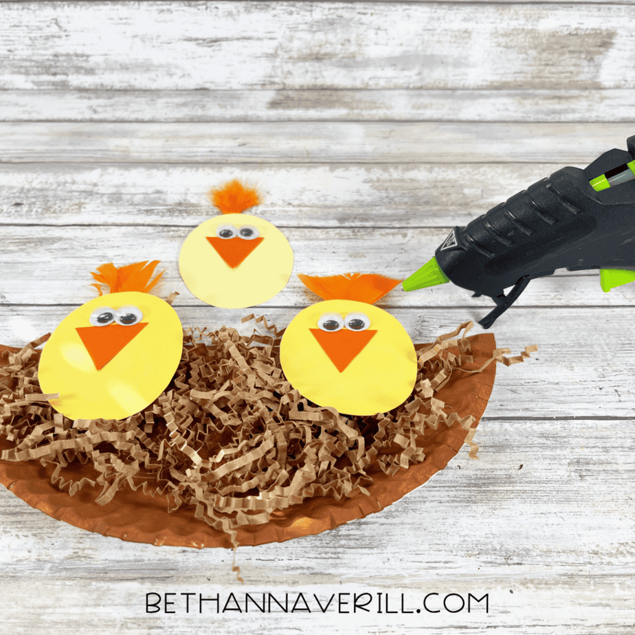 Three decorated yellow oval shapes are placed on top of the shredded paper on the plate half. A hot glue gun is nearby.