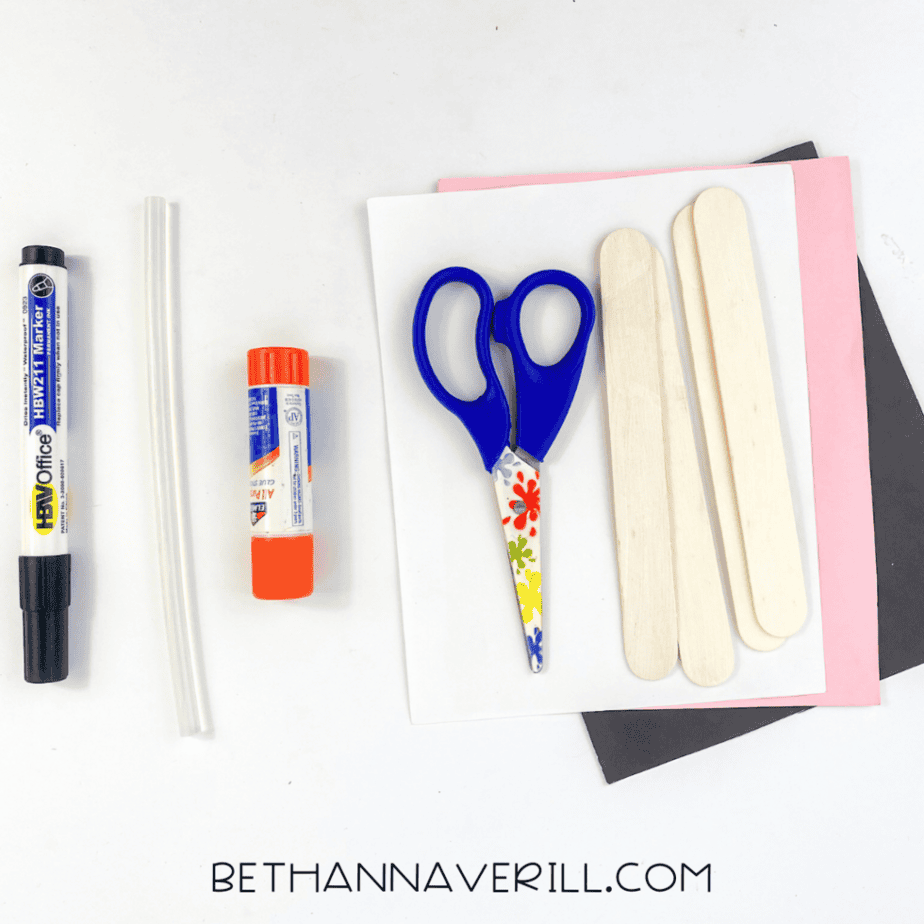 A black marker, two clear glue sticks, a glue stick container, scissors, several wooden craft sticks, and sheets of white, pink, and black paper are arranged on a white surface.