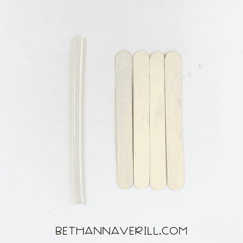 Four wooden craft sticks are lined up vertically side by side. Two clear glue sticks are placed to the left.