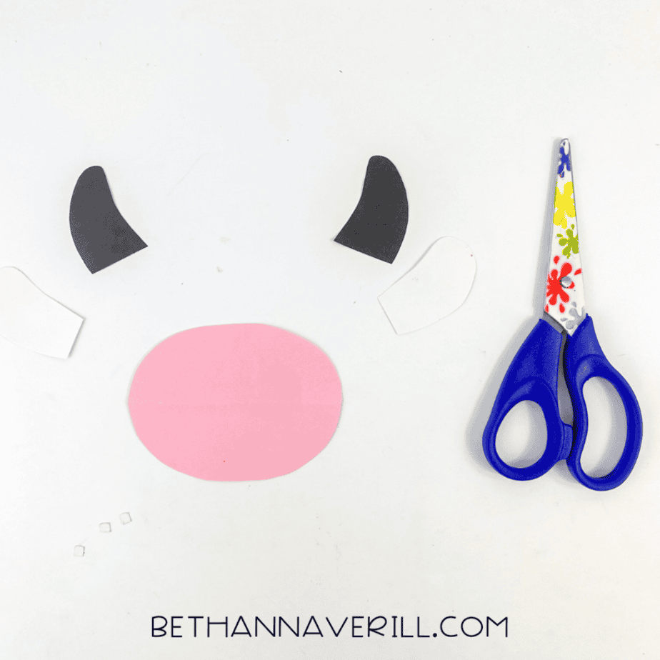 Cut paper shapes are laid out: a large pink oval, two black curved shapes, and two white shapes. A pair of scissors is on the right.