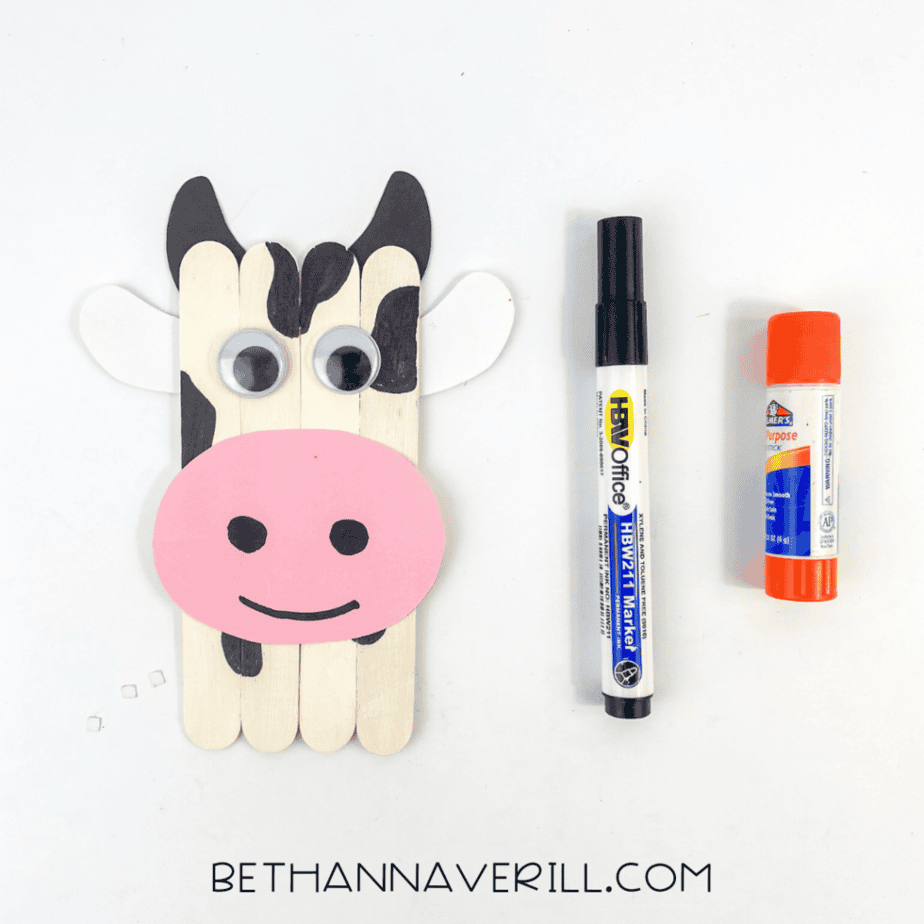 The wooden sticks are assembled vertically with added elements: googly eyes, a pink oval shape attached near the bottom, white shapes on the sides, black shapes at the top, and dark patches on the sticks. A marker and glue stick are placed to the right.